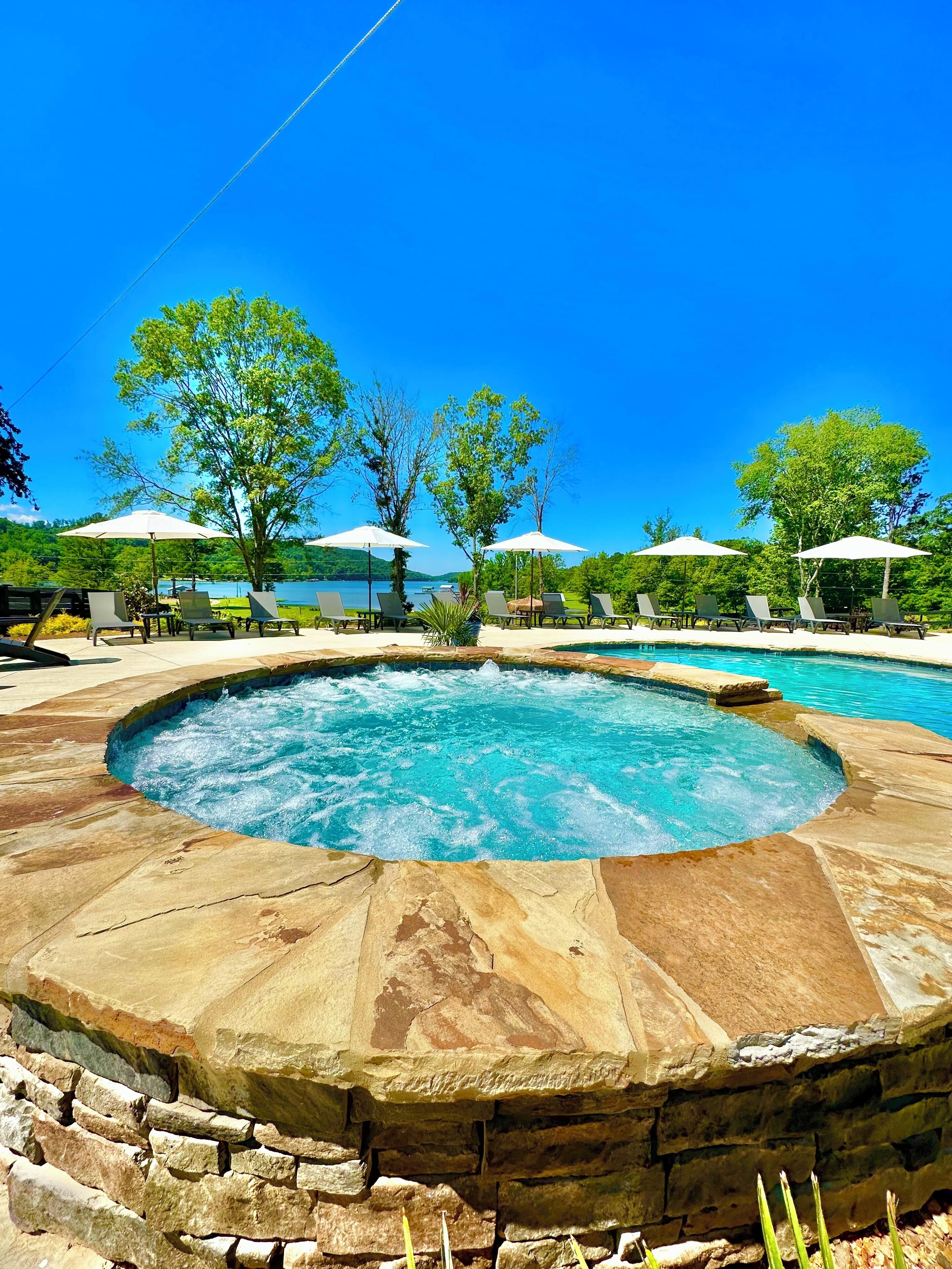 A circular hot tub surrounded by lounge chairs and umbrellas, with a view of a lake and trees in the background.