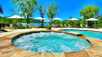 A circular hot tub surrounded by lounge chairs and umbrellas, with a view of a lake and trees in the background.
