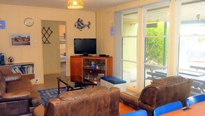 A living room with a brown leather sofa, a glass coffee table, and a TV on a wooden stand. There is a bookshelf with books and decorative items, and a dining area with a wooden table and blue chairs. - Gunnadoo, Moonta Bay (Moonta Bay)