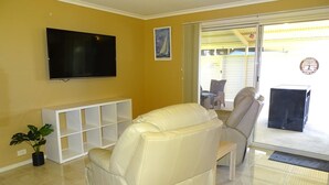 Ein Wohnzimmer mit einer Couch, einem Fernseher, einer weißen Regaleinheit und einer Glastür, die zu einem Außenbereich führt.