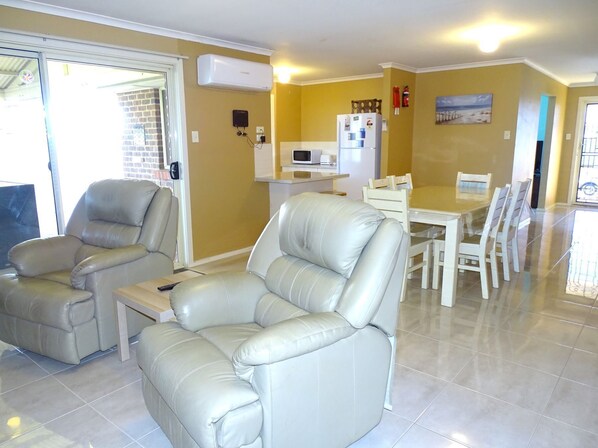 A living room with a sofa, a dining table with chairs, a kitchen area with appliances, and a sliding glass door leading to a balcony.