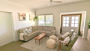 A living room with a beige sectional sofa, a wooden coffee table, and two ottomans. There are two windows with blinds, a ceiling fan, and a door with a window.