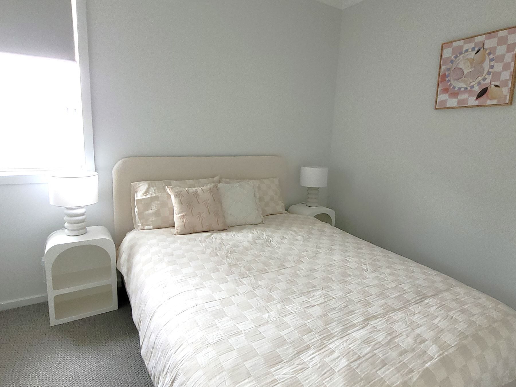 Un dormitorio ordenado con una cama, dos mesitas de noche con lámparas, una ventana y un cuadro colgado en la pared.