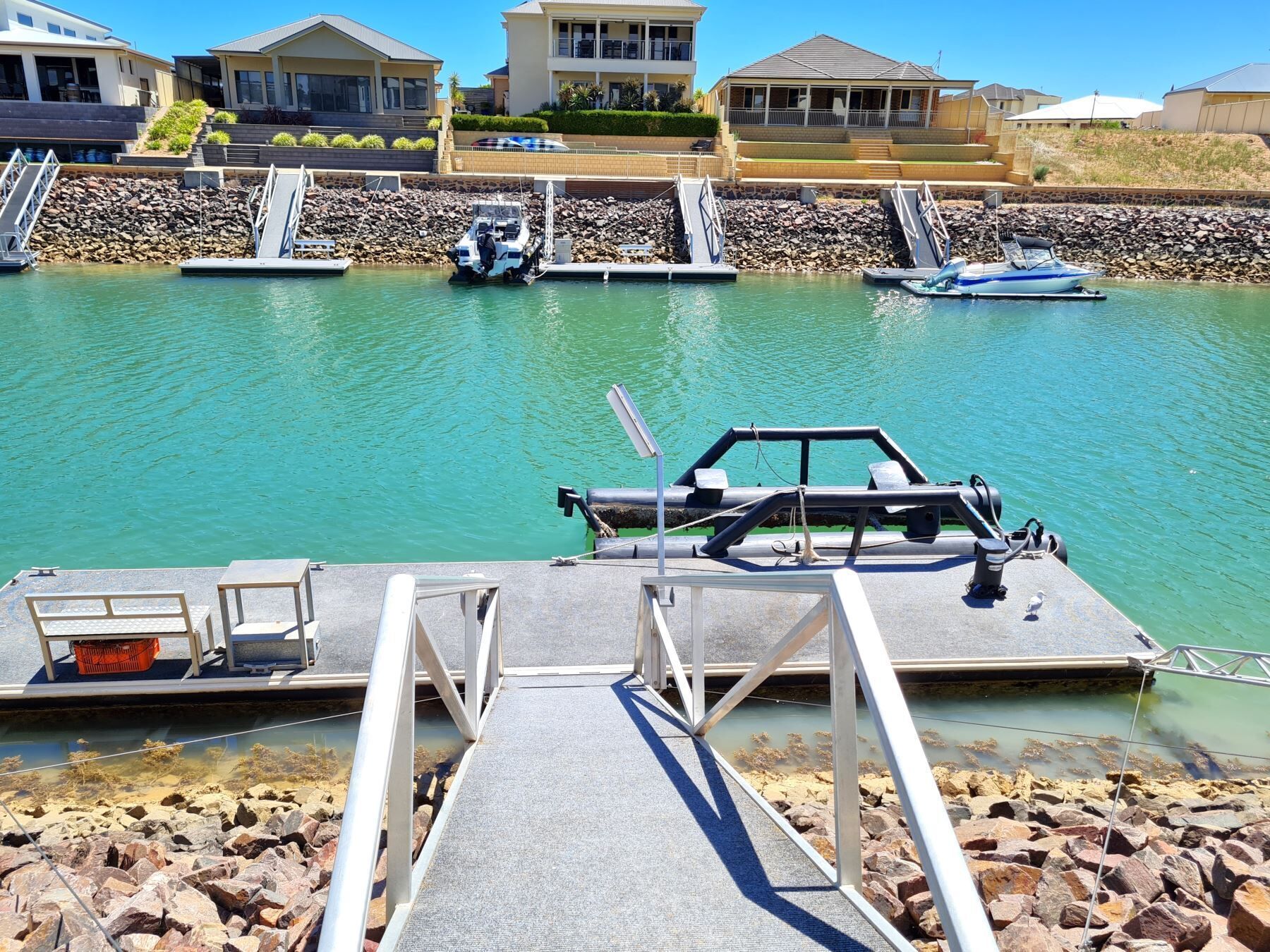 A marina with boats docked, a concrete dock with steps leading into the water, and houses in the background.