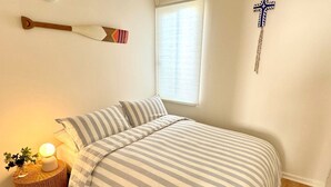 Un lit bien fait, avec une literie à rayures, une lampe de chevet, une petite table d’appoint en osier ornée d’une plante et un crucifix fixé au mur.