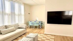 Un salon comprenant un canapé, des poufs, une table avec des chaises et un téléviseur fixé au mur.
