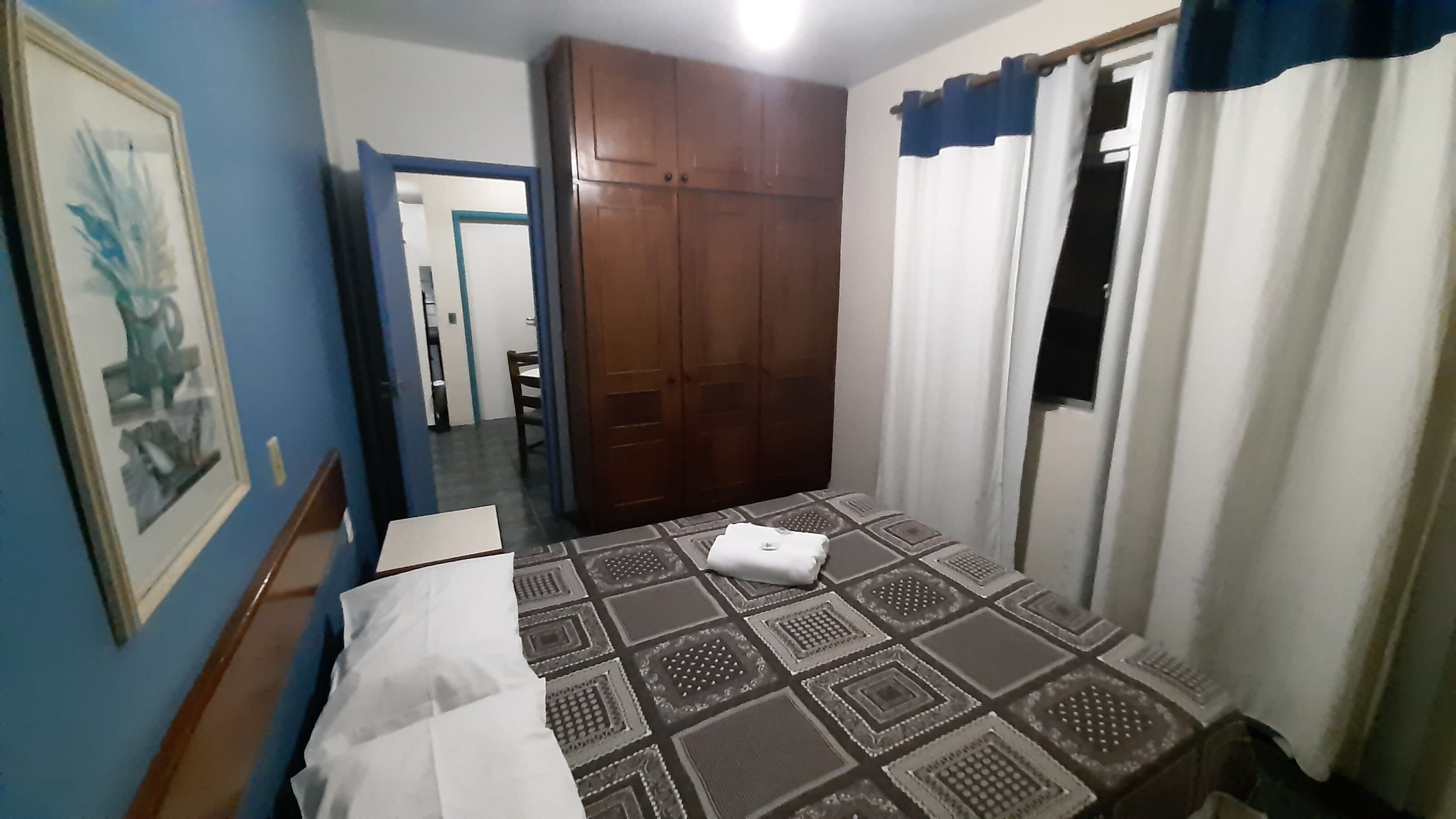 Un dormitorio con cama, armario, un cuadro enmarcado en la pared y una ventana con cortinas.