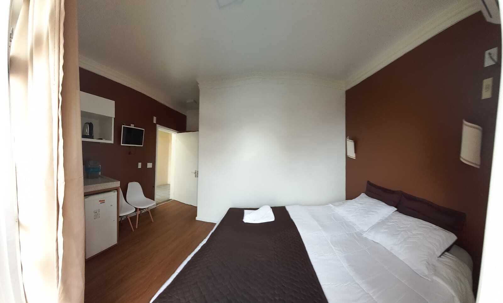 A hotel room with a bed, a desk with a chair, a small refrigerator, and a TV.