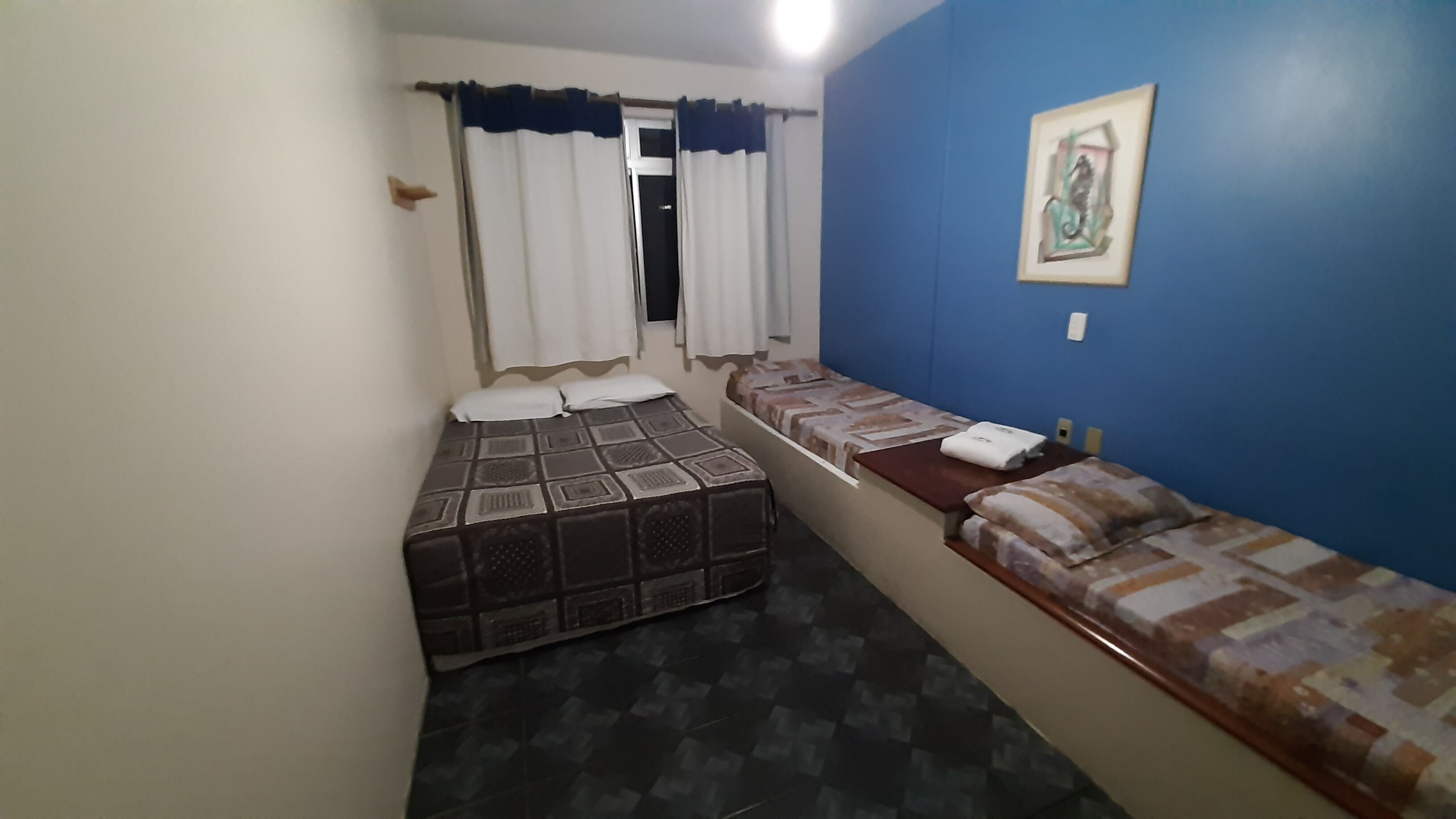 A small, narrow room with two beds, a window with curtains, a painting on the wall, and a tiled floor.
