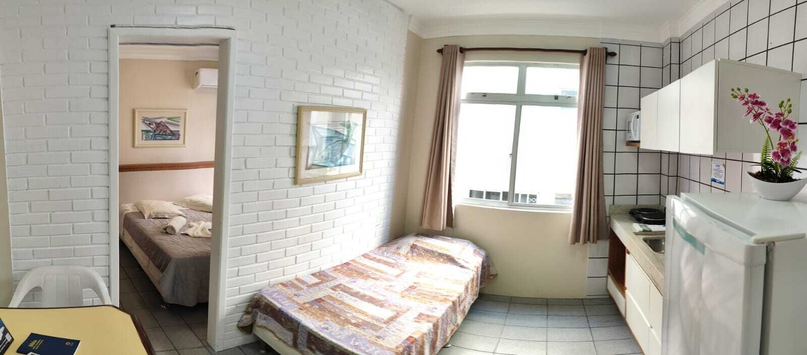 A compact room with a bed, a small table, a window with curtains, and a kitchenette.