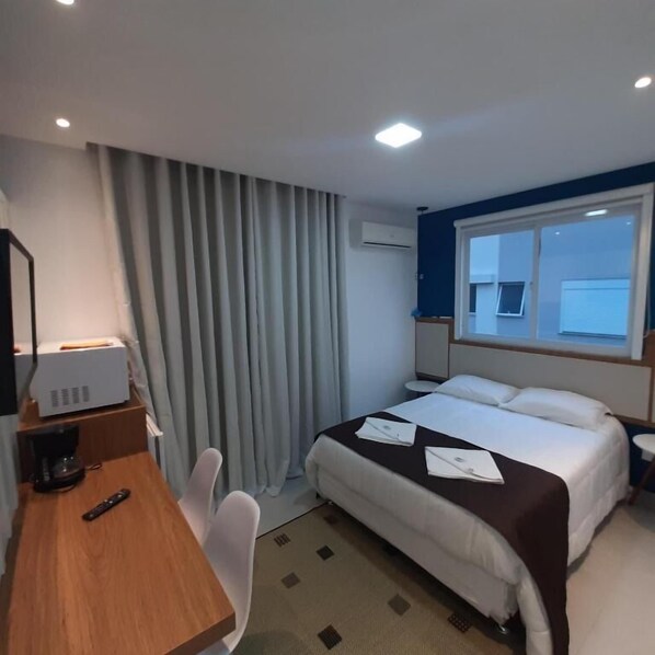 A hotel room with a bed, a desk with a computer, a chair, a window, and an air conditioning unit.