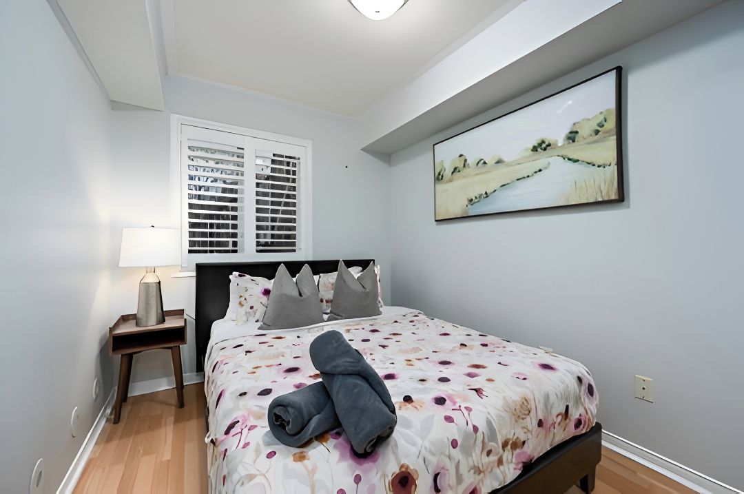 A bedroom with a bed, a nightstand, a lamp, a window with blinds, and a painting on the wall.
