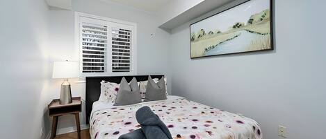 A bedroom with a bed, a nightstand, a lamp, a window with blinds, and a painting on the wall.