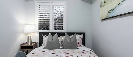 A bedroom with a bed, a nightstand, a lamp, a chair, and a window with blinds.
