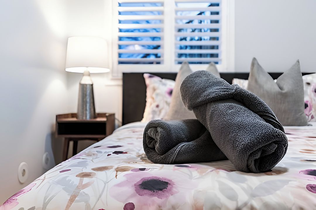 A neatly made bed with a floral-patterned bedspread, a lamp on a bedside table, and a window with blinds in the background.