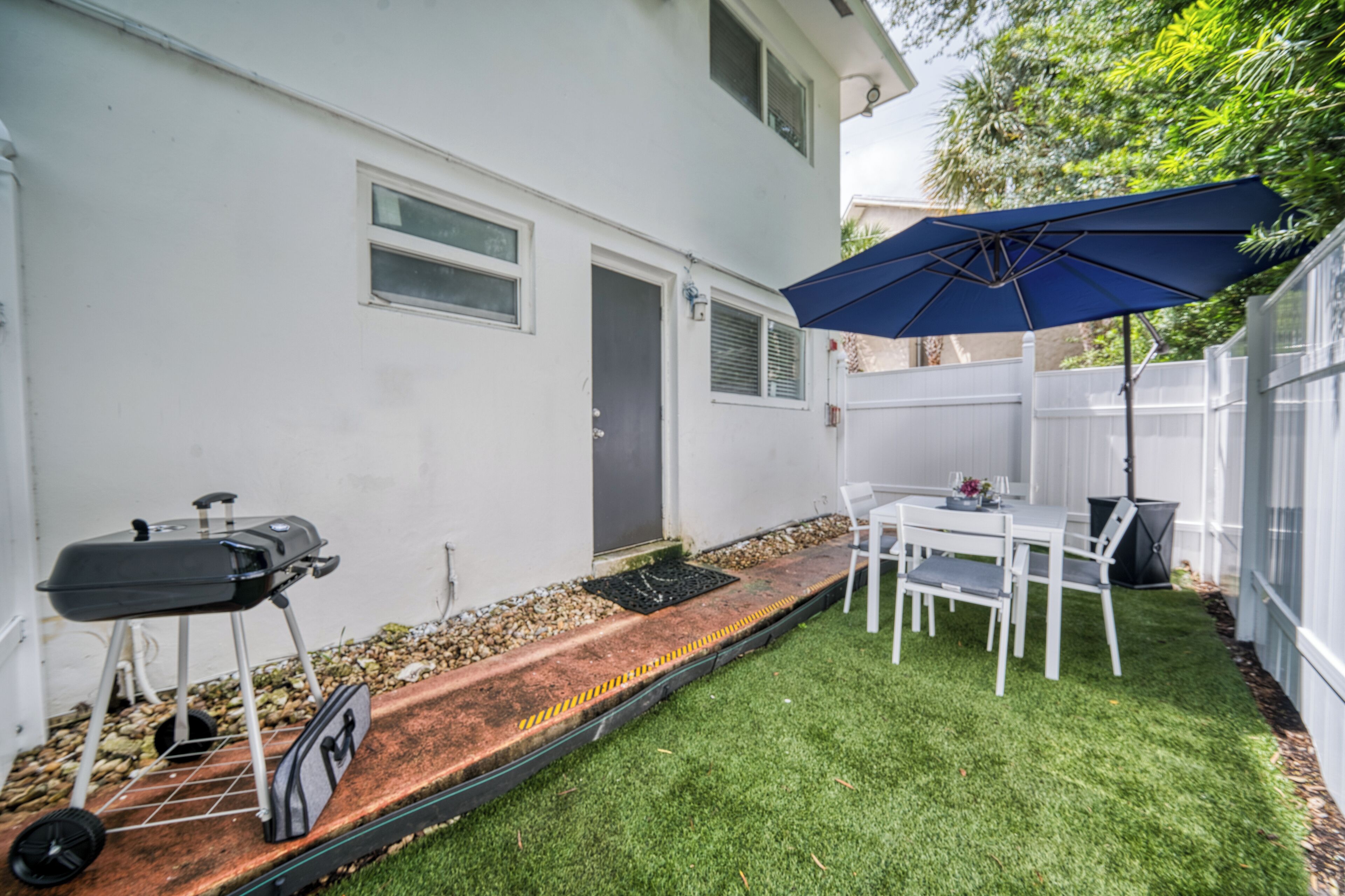 A backyard with a grill, a table with chairs, and a blue umbrella.