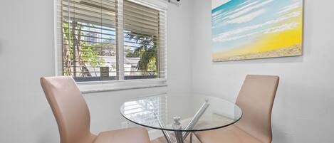 Habitación con una mesa de vidrio, dos sillones de cuero, una ventana con persianas y un cuadro de playa en la pared.