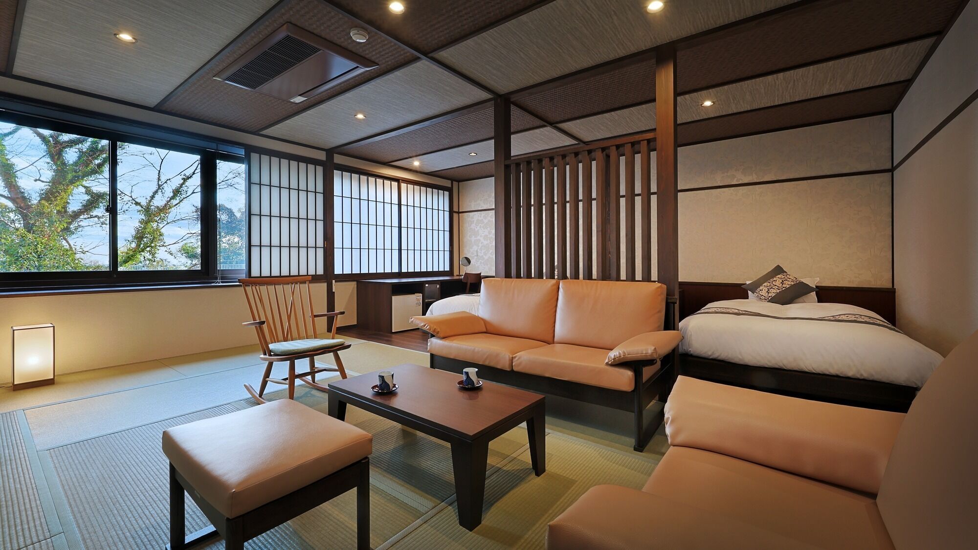 A traditional Japanese-style room with tatami flooring, a low wooden table, a sofa, a bed, and a view of trees outside.