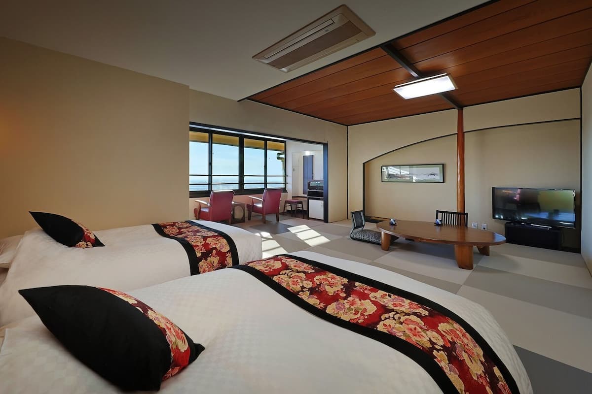 A hotel room with two beds, a dining table, chairs, a TV, and a view of the sea.