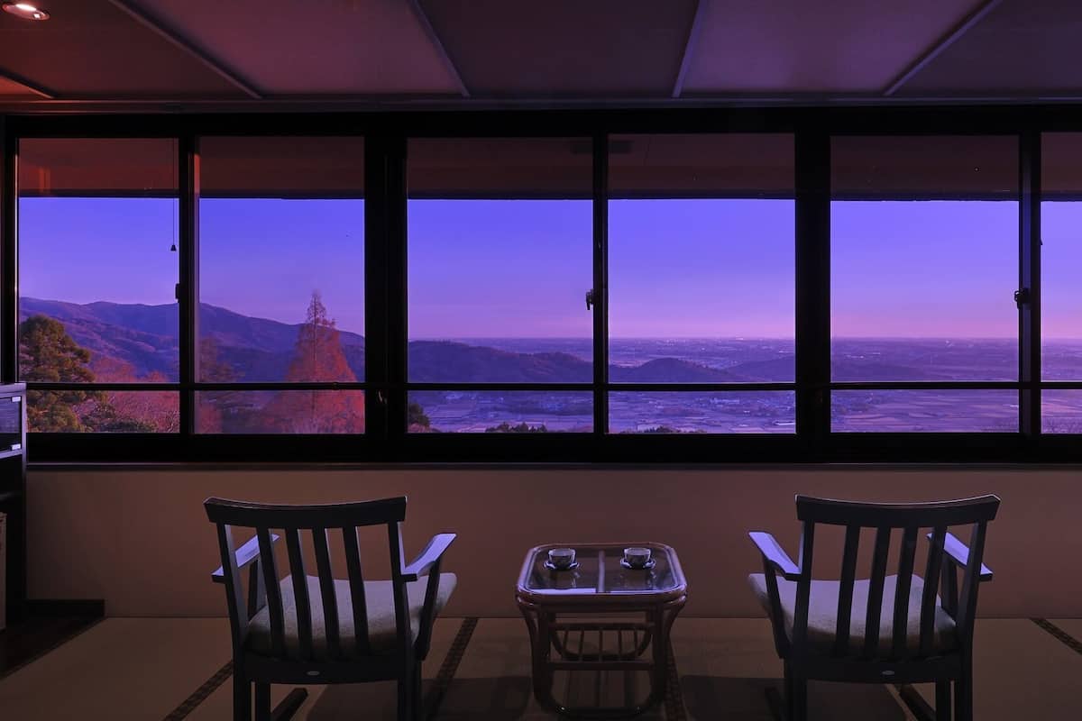 A room with a large window overlooking a mountainous landscape. There are two chairs and a small table in the room.