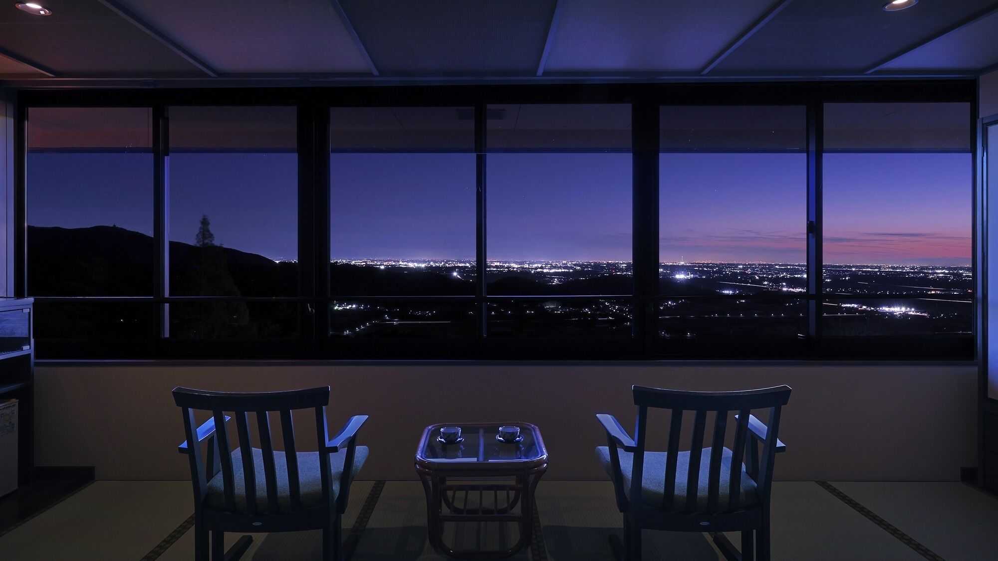 A room with a large window overlooking a cityscape at dusk. There are two chairs and a small table in the foreground.