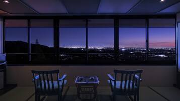 A room with a large window overlooking a cityscape at dusk. There are two chairs and a small table in the foreground.