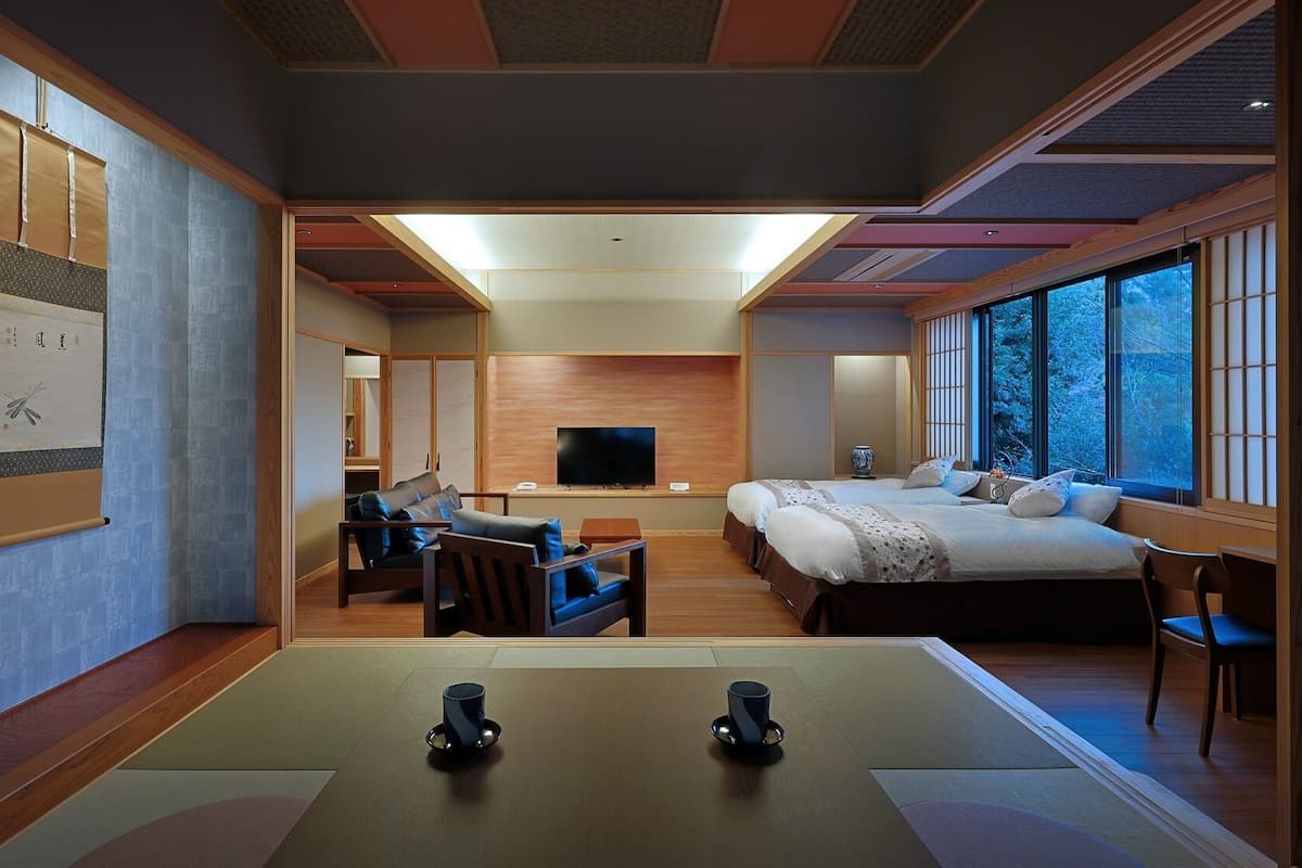 A hotel room with a large bed, a TV, a desk, and a view of trees through the window.