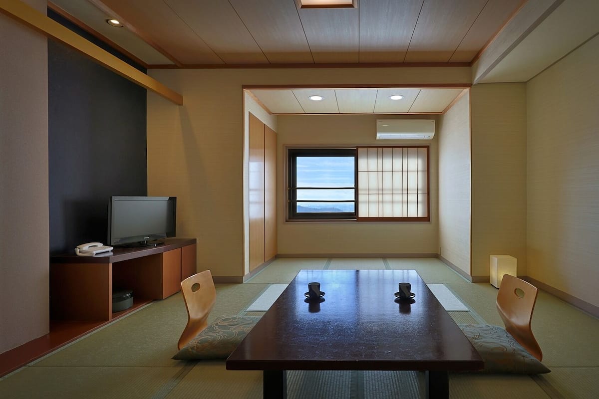 A traditional Japanese room with tatami flooring, a low table with cushions, a television, and a window with a view of the mountains.