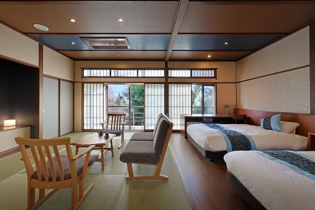 A traditional Japanese-style hotel room with tatami flooring, a low table with a cushion, a sofa, and two beds with pillows.