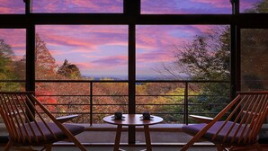 Un balcon avec deux chaises en bois et une petite table, d’où l’on peut admirer un paysage magnifique, avec les arbres aux couleurs automnales et le ciel au coucher du soleil.