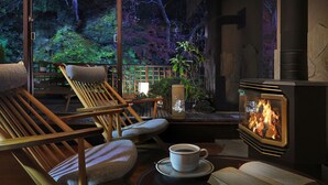 Une pièce chaleureuse avec une cheminée, des chaises en bois, une table avec une tasse, et une vue sur la verdure à l’extérieur.
