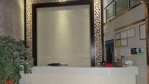 A reception desk with a patterned wallpaper backdrop, a potted plant to the left, and various notices on the wall. - Senli Hotel (Sanmen)