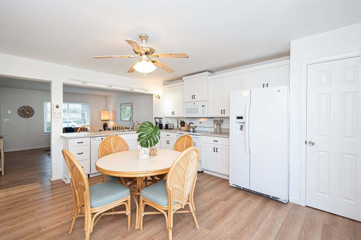 A modern kitchen with white cabinets, a dining area with wicker chairs, a ceiling fan, and a refrigerator.