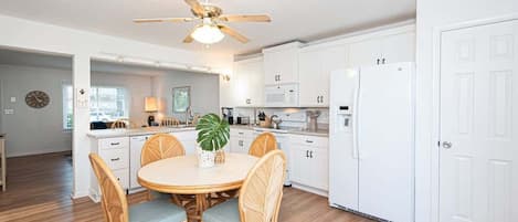 A modern kitchen with white cabinets, a dining area with wicker chairs, a ceiling fan, and a refrigerator.