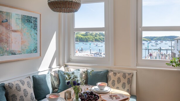 A dining area with a view of boats on the water, a wicker chandelier, and a table set with plates, glasses, and a bowl of fruit.