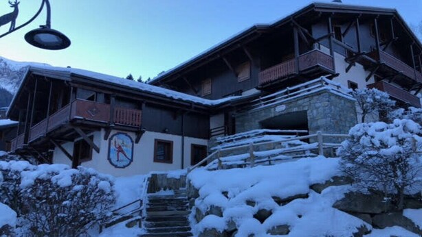 Ein traditionelles Chalet mit einer Steintreppe, die zu einer Holzdiele führt, umgeben von schneebedeckten Bäumen und einem Berg im Hintergrund.