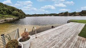 Un muelle de madera se extiende sobre un lago tranquilo, con una zona de césped y una cerca de malla metálica en un lado.