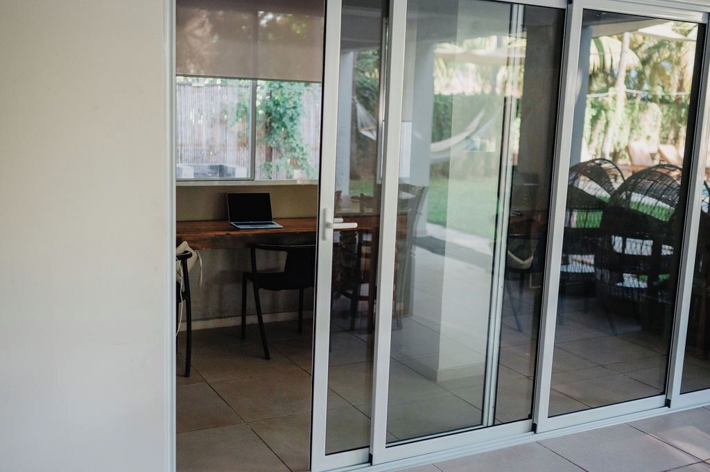 A room with a sliding glass door leading to a patio area. There is a laptop on a desk, a chair, and a dining table with chairs.