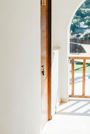 A wooden door with a brass handle, leading to a balcony with a white railing and a view of greenery.