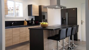 A modern kitchen with wooden cabinets, a central island with bar stools, a stainless steel refrigerator, and a window with a view of buildings. - Maison Loutina, Villa 5-10 min à Pied du Centre (L'Isle-sur-la-Sorgue)