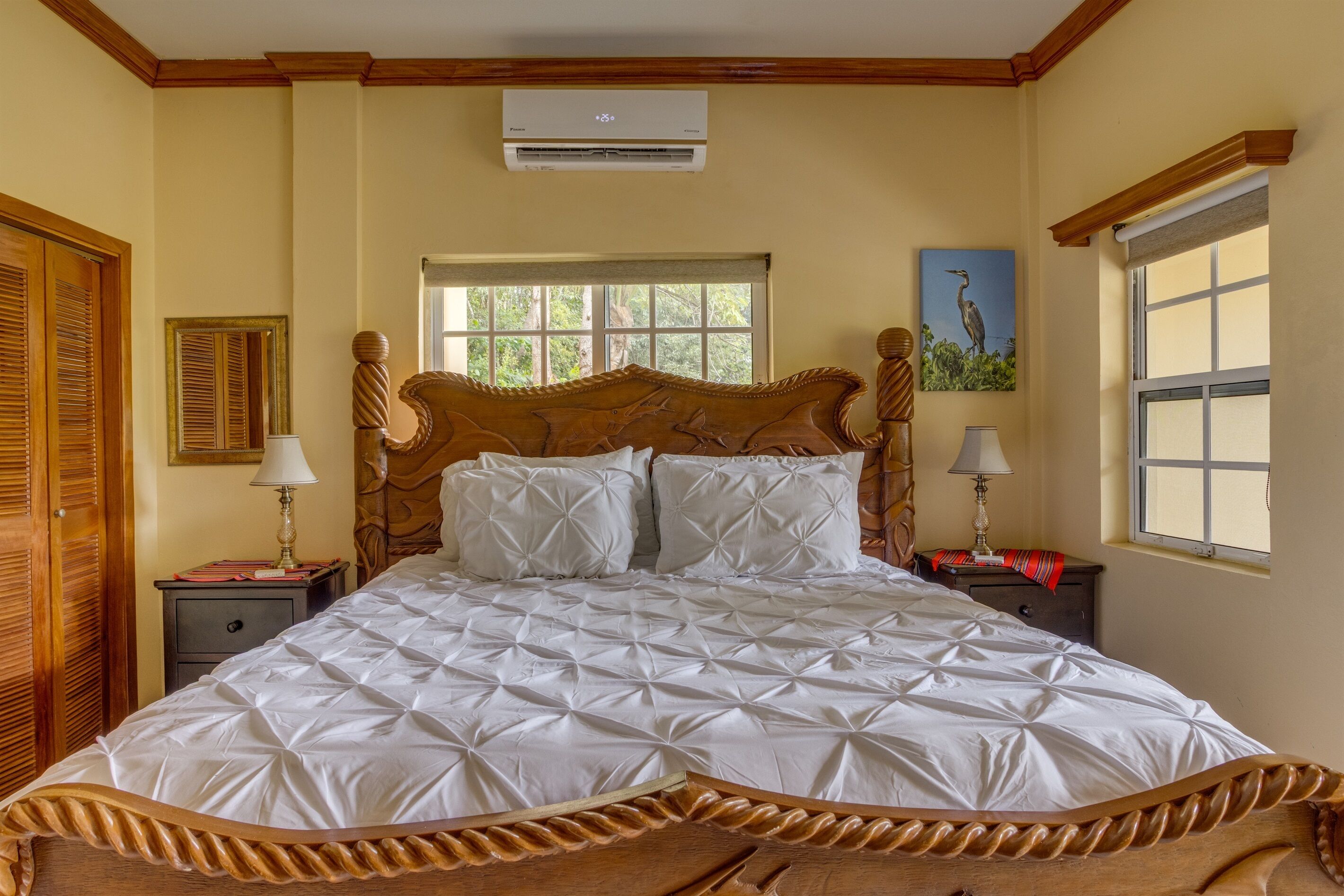 Un dormitorio con una cama grande, cabecera de madera, dos mesitas de noche con lámparas, una ventana con vistas a zonas verdes y un equipo de aire acondicionado en la pared.