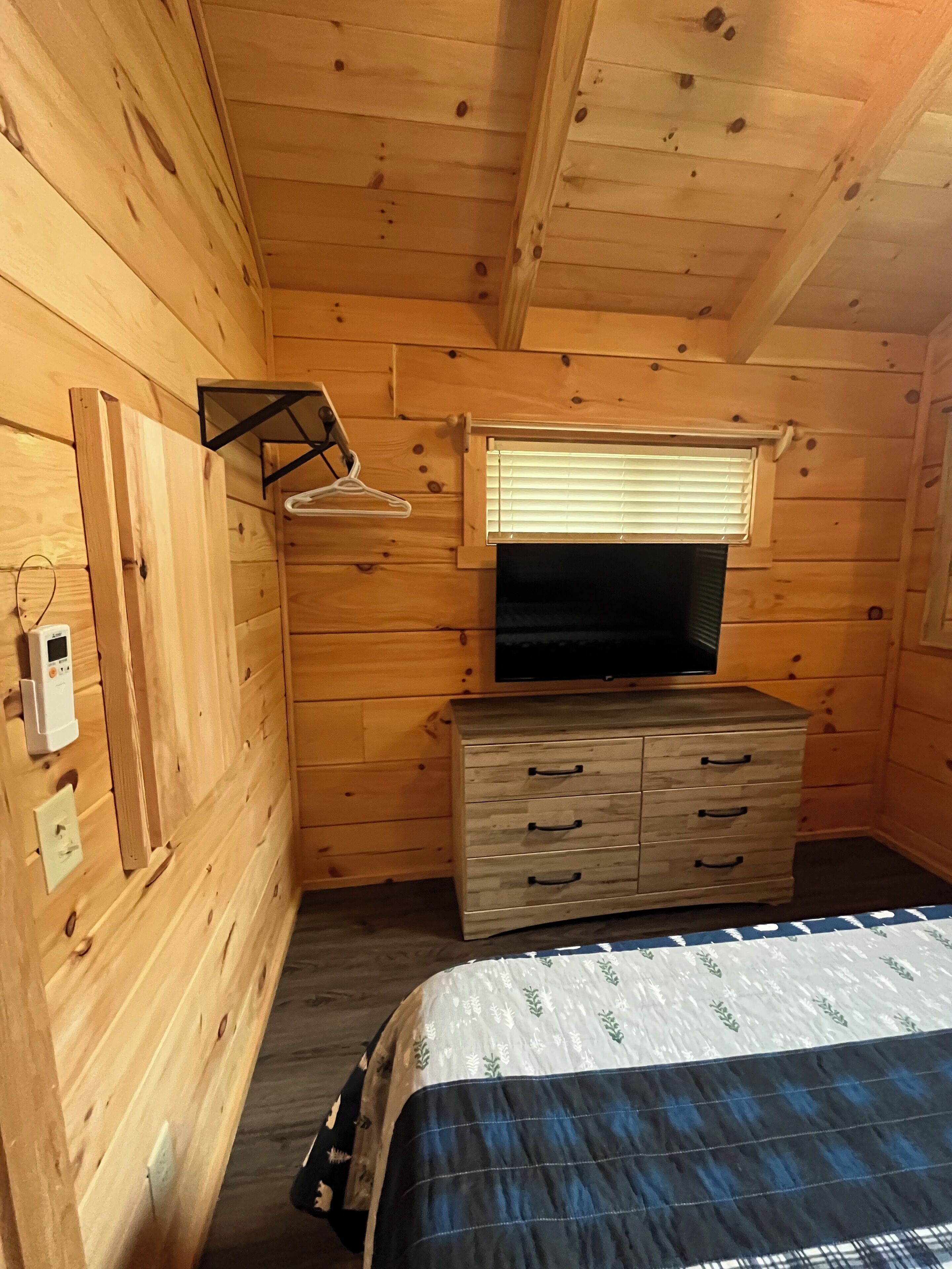 Une chambre dans un chalet en bois, avec un lit, une commode, un téléviseur et une fenêtre équipée de stores.