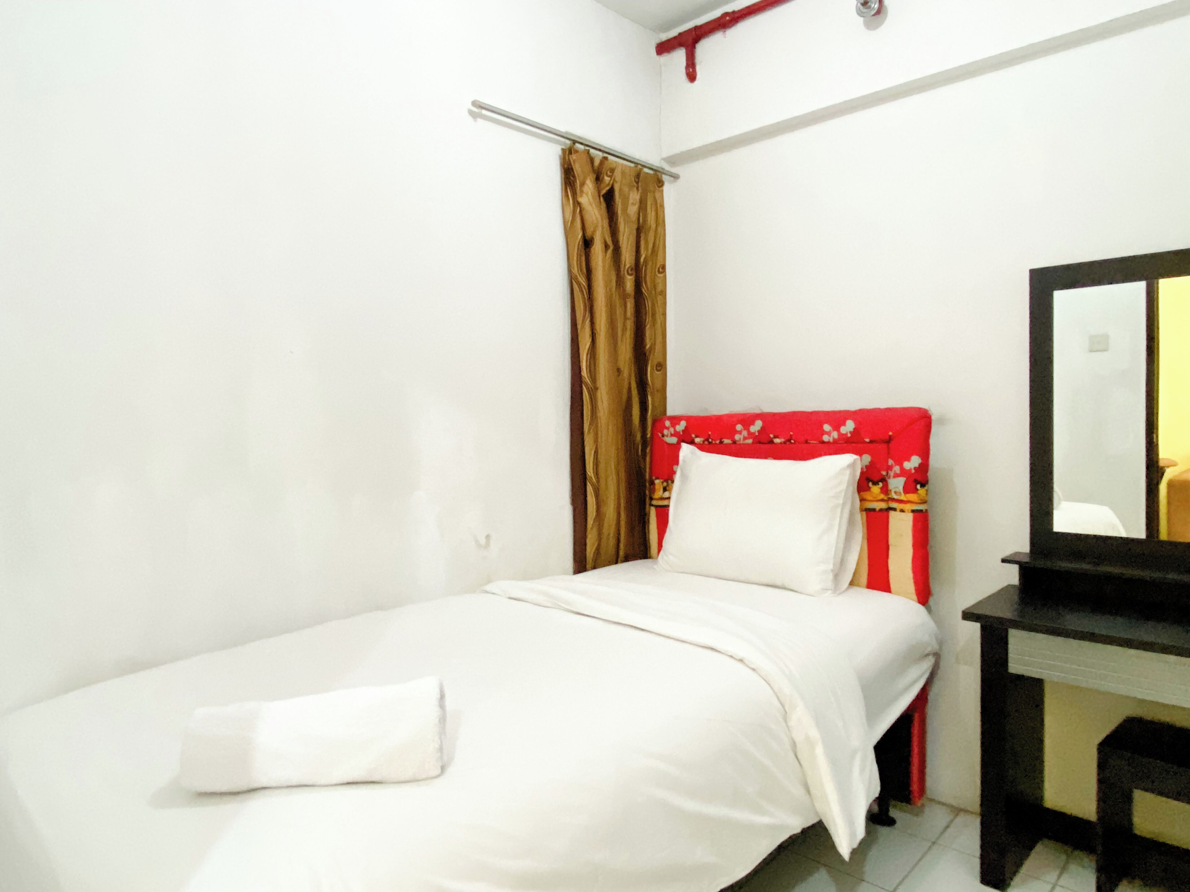 A small, neatly made bed with white linens and a white pillow, a red headboard with a floral pattern, a wooden nightstand with a mirror, and a brown curtain partially drawn.