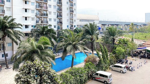 A high-rise building with multiple balconies, a swimming pool surrounded by palm trees, and a parking area with vehicles. - Warm And Cozy 2Br Mutiara Bekasi Apartment (Bekasi)