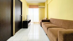 A living room with a brown sofa, a TV on a stand, and a door. - Warm And Cozy 2Br Mutiara Bekasi Apartment (Bekasi)