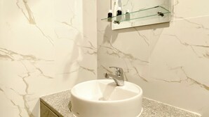 Un baño con pared de mármol, un lavamanos con grifo y un estante de vidrio sobre el lavamanos que contiene artículos de aseo.