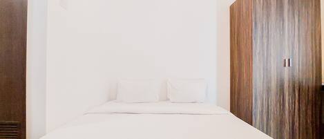 A bedroom with a bed, white bedding, a wooden wardrobe, and a white wall.