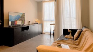 A modern living room with a sofa, a TV on a black cabinet, and a window with sheer curtains. - Spacious 1Br At Tokyo Riverside Pik 2 Apartment (Teluknaga)