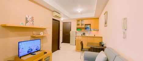 A compact living space with a TV on a wooden stand, a grey sofa, a kitchen area with a microwave, and a small dining table.