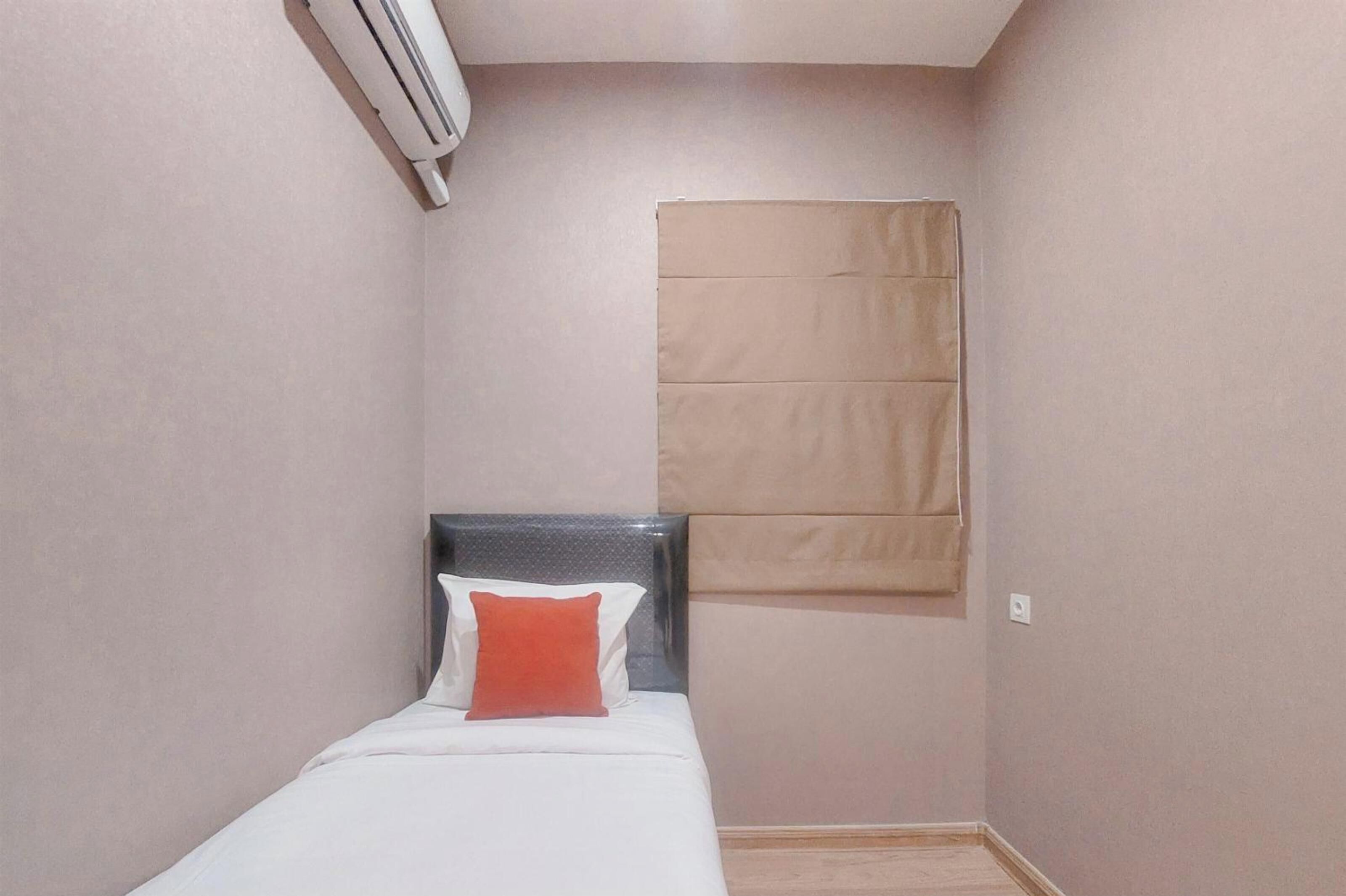 Une petite chambre avec un lit simple, une tête de lit, un oreiller rouge et une fenêtre avec un store beige.